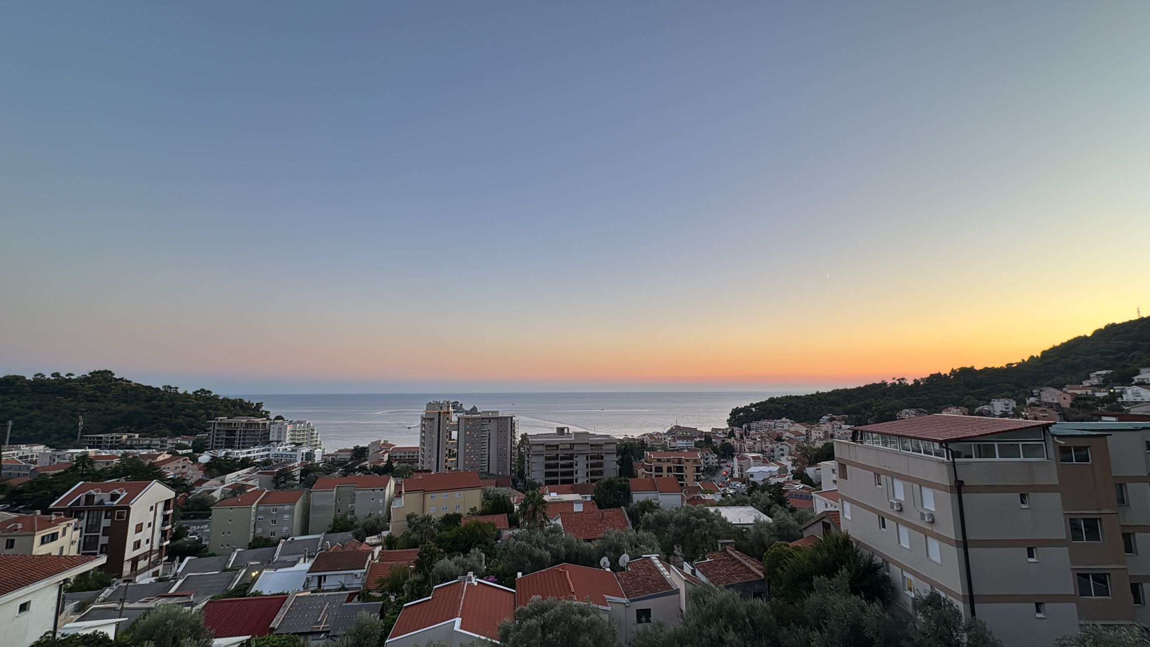 Petrovac u sumrak: krovovi i zgrade spuštaju se ka moru, nad horizontom narandžasti sjaj poslije zalaska sunca